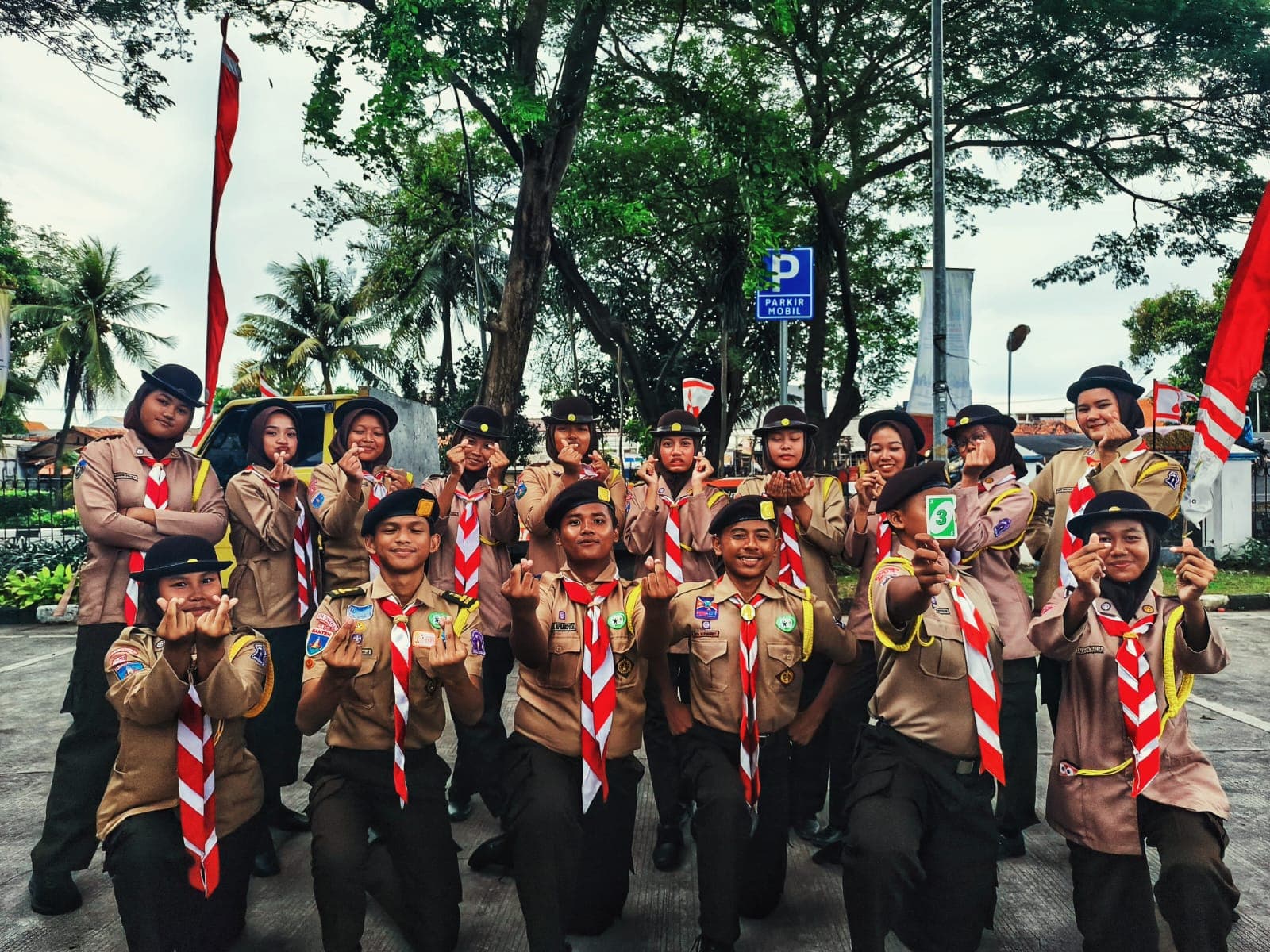 Sekelompok anggota Pramuka berseragam lengkap berpose dengan simbol hati di jari tangan di luar ruangan.