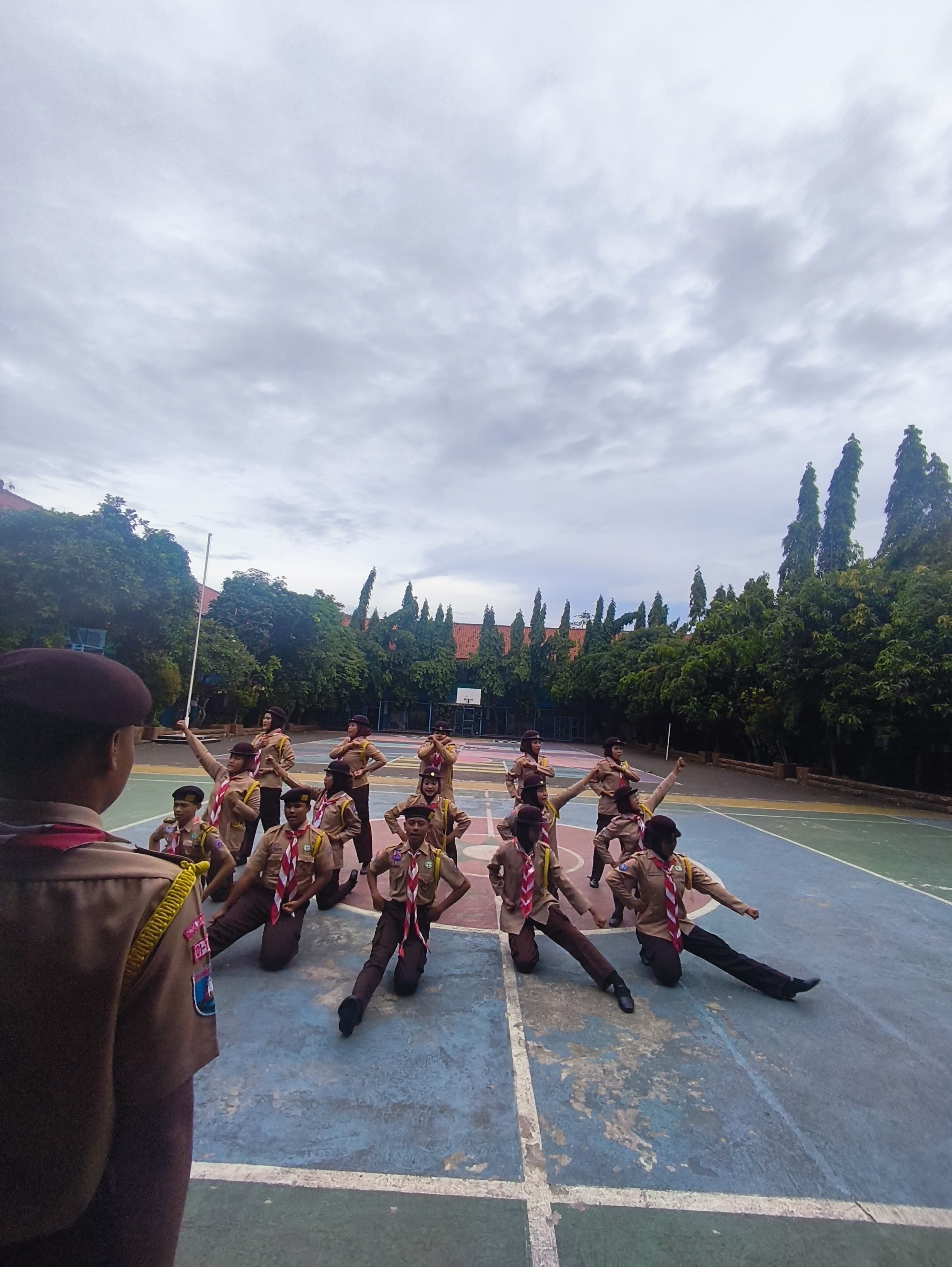 Sekelompok anggota Pramuka melakukan gerakan tari sinkron di lapangan basket di bawah langit mendung.