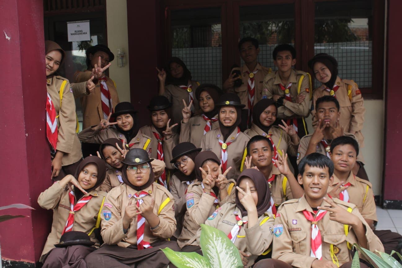 Sekelompok siswa Indonesia berseragam pramuka cokelat berpose ceria dengan berbagai gaya di depan sekolah.