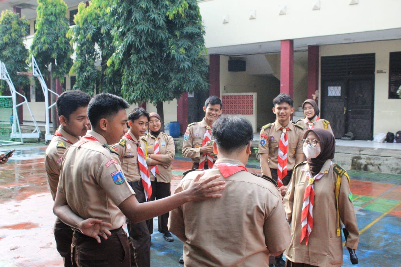 Sekelompok anggota Pramuka berseragam cokelat sedang berkumpul dan berinteraksi dengan ceria di lapangan sekolah.
