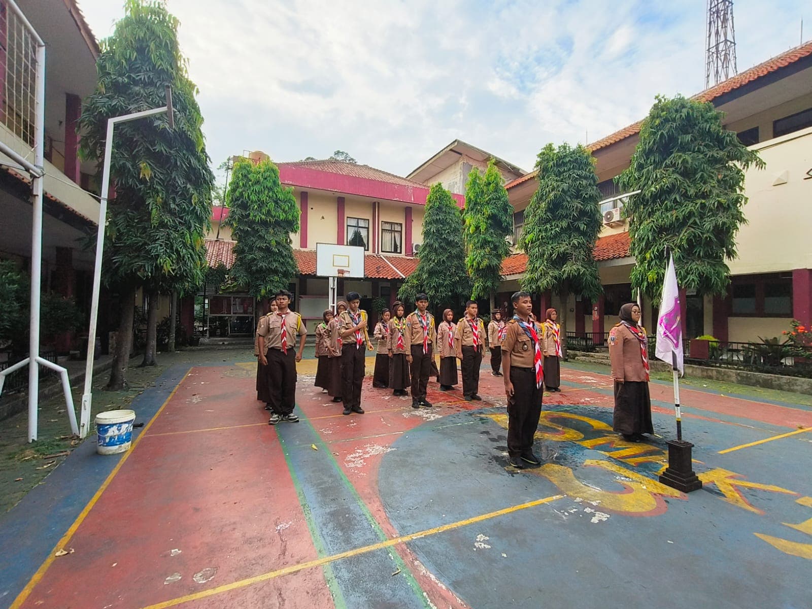 Sekelompok siswa berseragam Pramuka berdiri dalam formasi di lapangan sekolah dengan latar belakang gedung.
