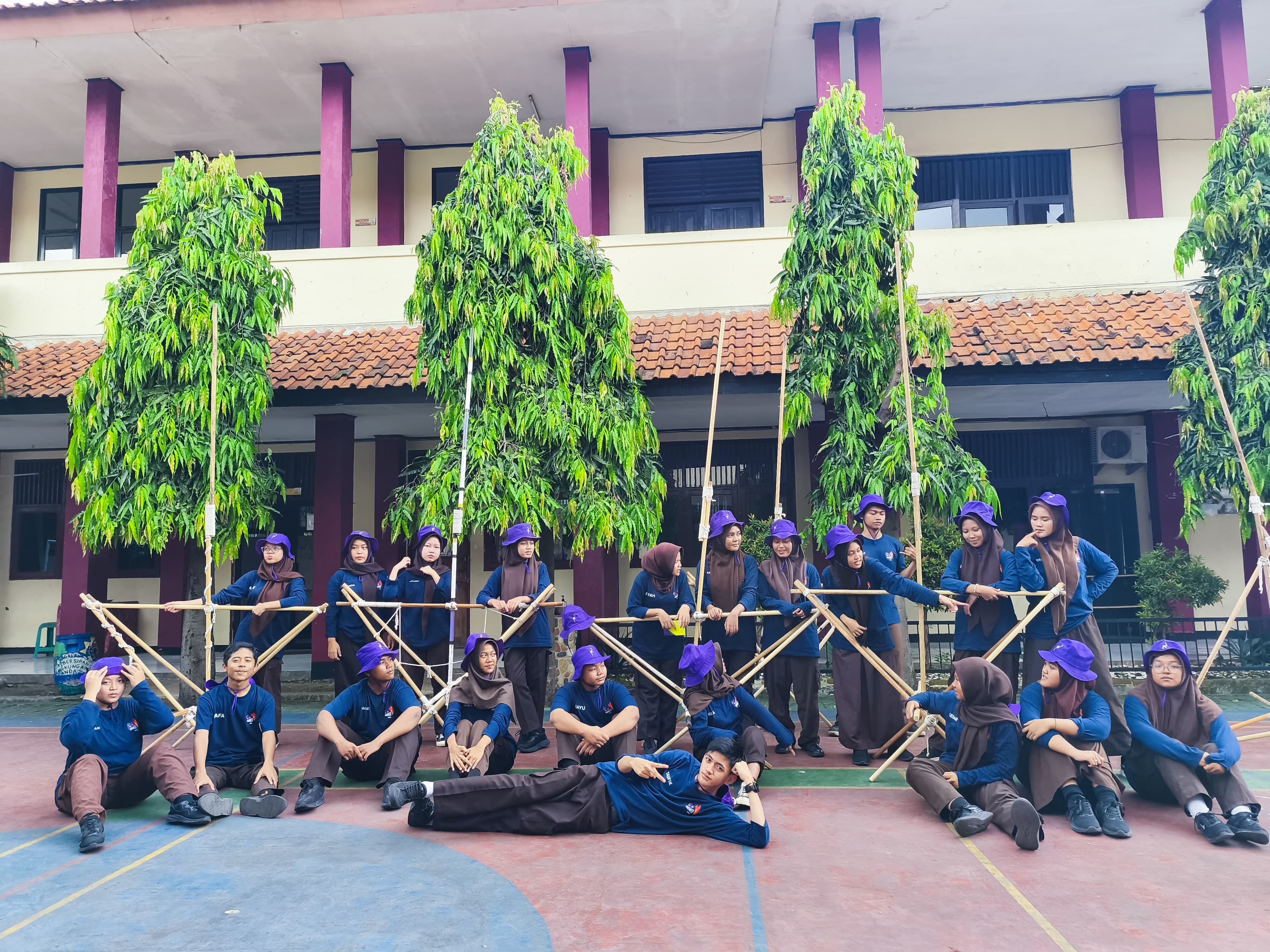 Sekelompok siswa berseragam biru dan topi ungu berpose dengan struktur bambu di halaman sekolah.
