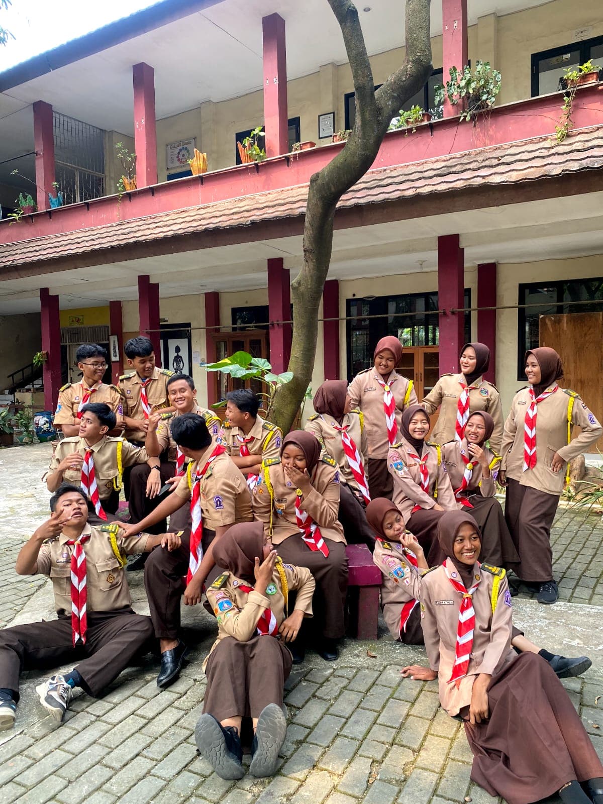 Sekelompok siswa berseragam Pramuka berpose ceria dan tertawa bersama di halaman sebuah sekolah.
