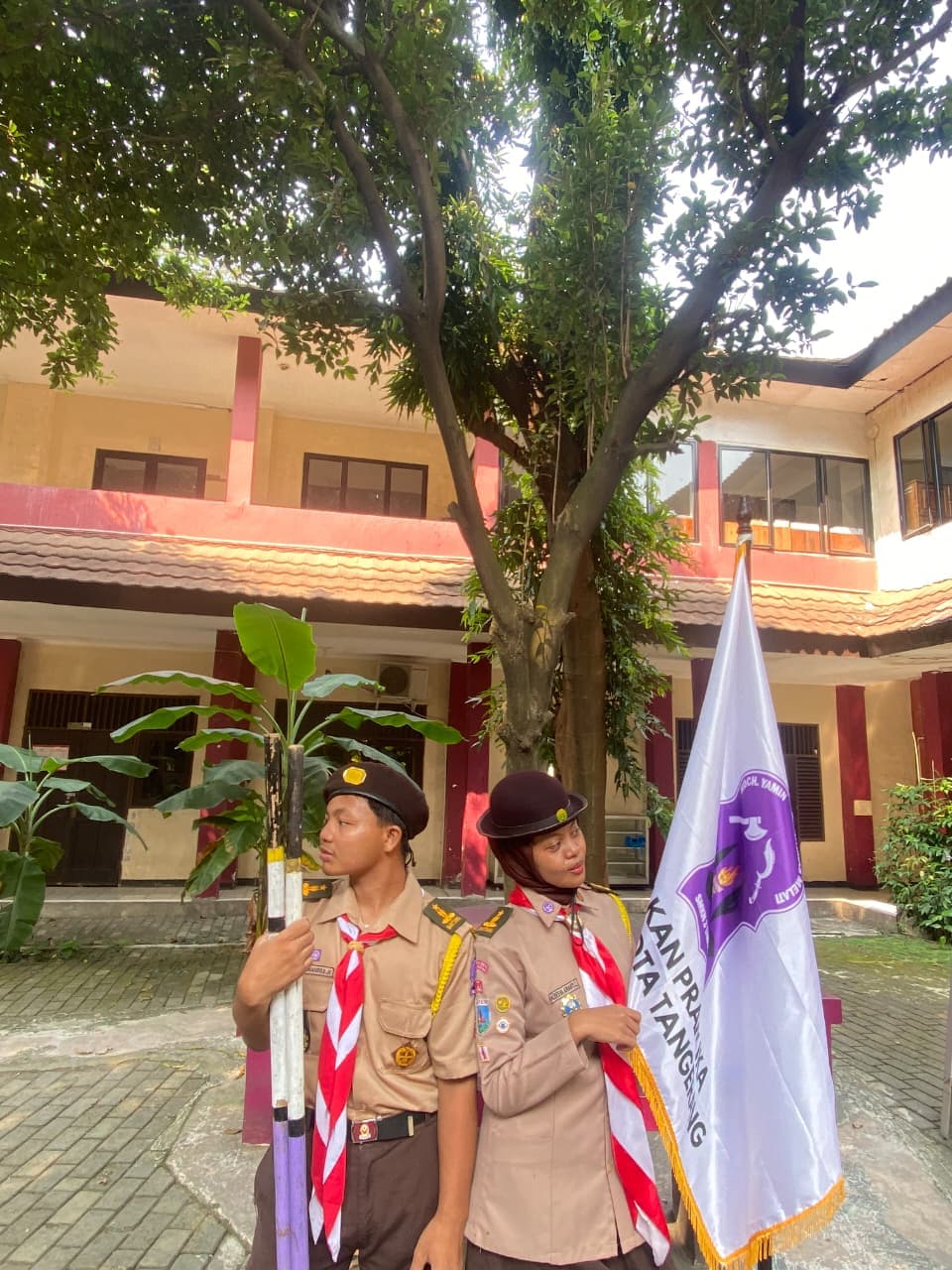 Dua anggota pramuka berseragam lengkap berpose di halaman sekolah membawa bendera dan tongkat kayu.