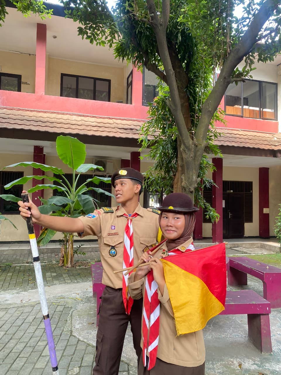 Dua anggota Pramuka berseragam lengkap berpose membawa tongkat dan bendera semaphore di halaman sekolah.