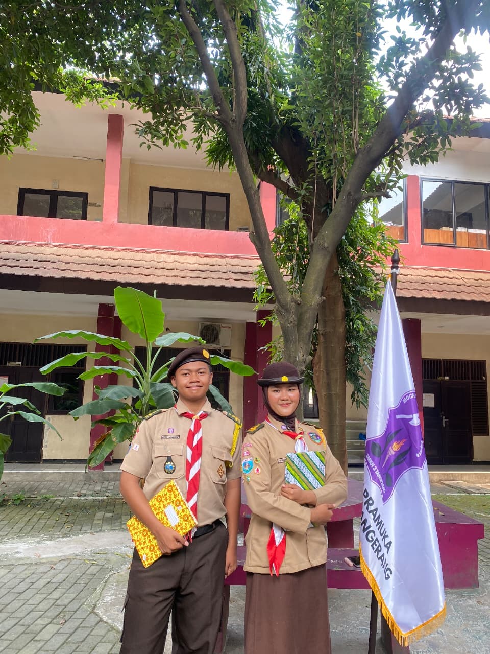 Dua siswa berseragam Pramuka berpose di depan gedung sekolah sambil memegang buku dan bendera.