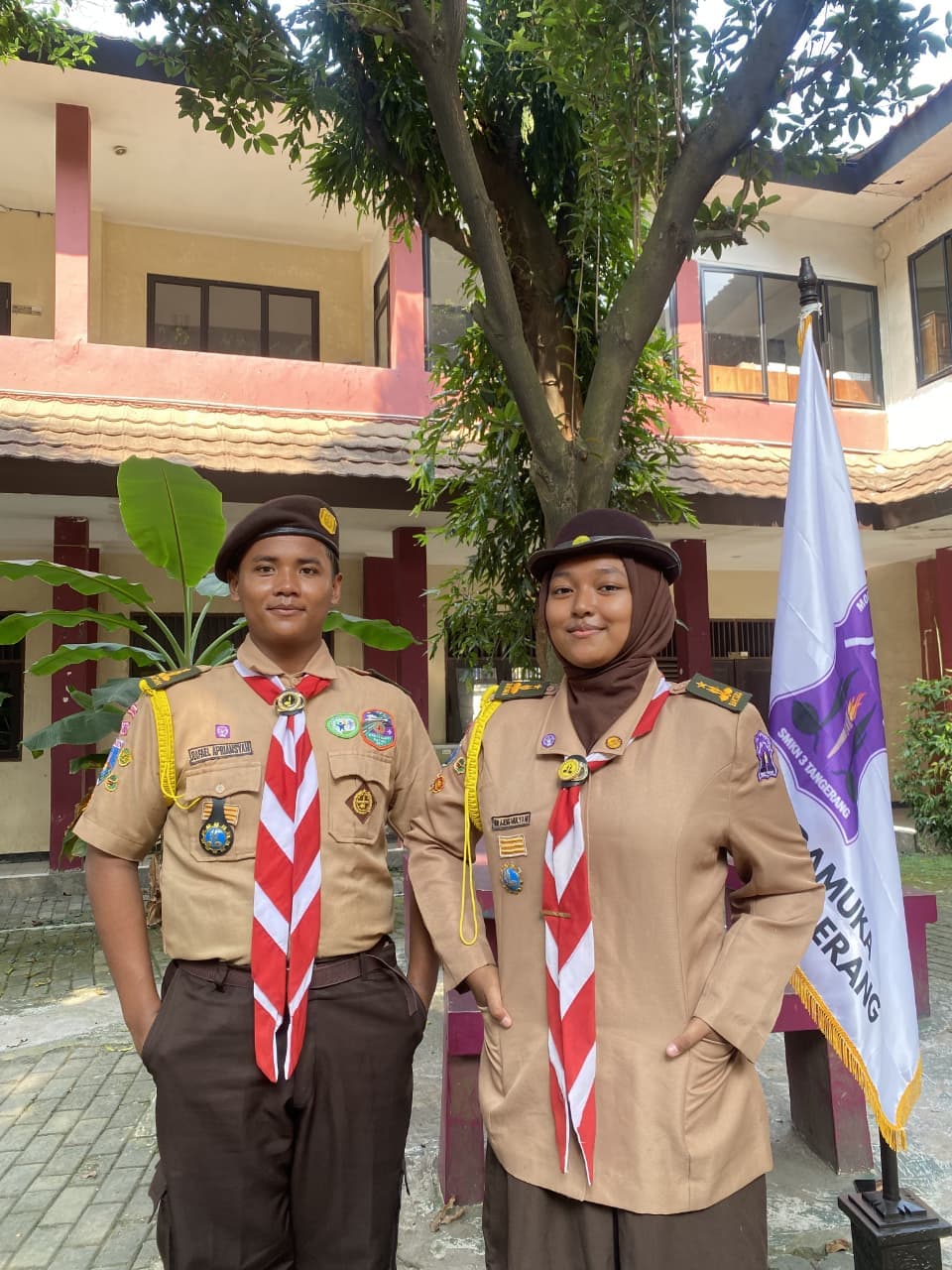 Dua anggota Pramuka berseragam lengkap berpose di depan gedung sekolah dengan latar pohon hijau.