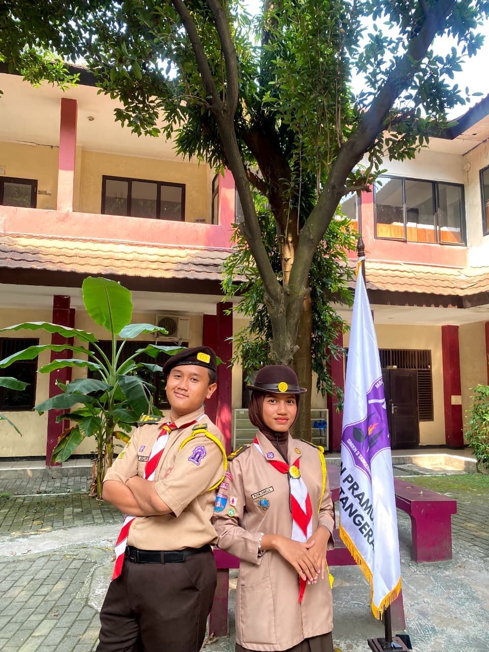 Dua anggota Pramuka berpose di depan gedung sekolah dengan latar belakang pohon dan bendera.