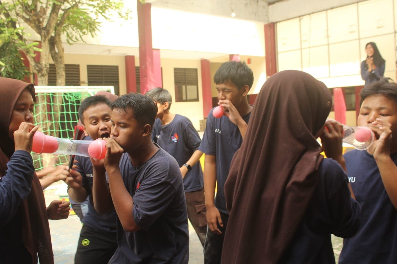 Sekelompok siswa berseragam biru sedang berlomba meniup balon merah muda melalui gelas plastik transparan.