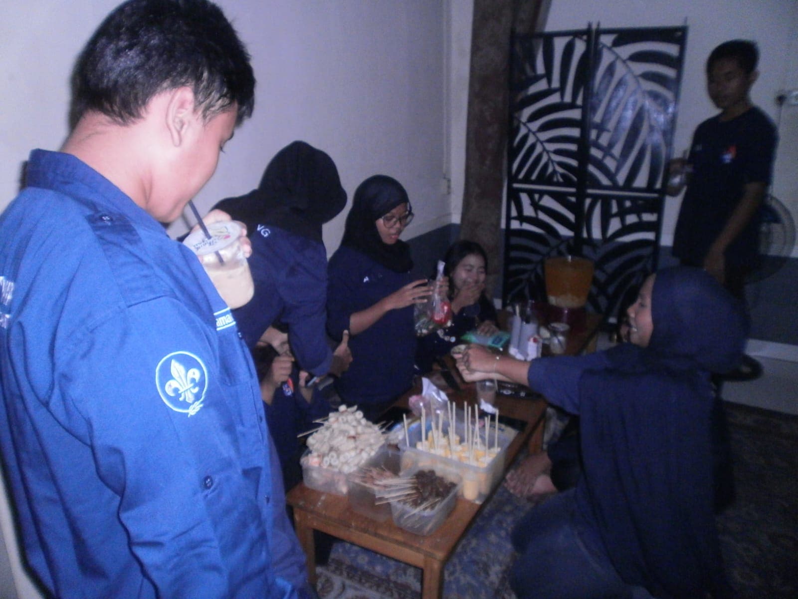 Sekelompok pemuda berseragam biru berkumpul di meja makan kecil sambil menikmati berbagai jajanan tusuk.