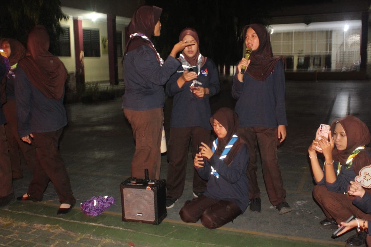 Siswi pramuka berhijab berkumpul di malam hari, satu orang berbicara menggunakan mikrofon dekat pengeras suara.