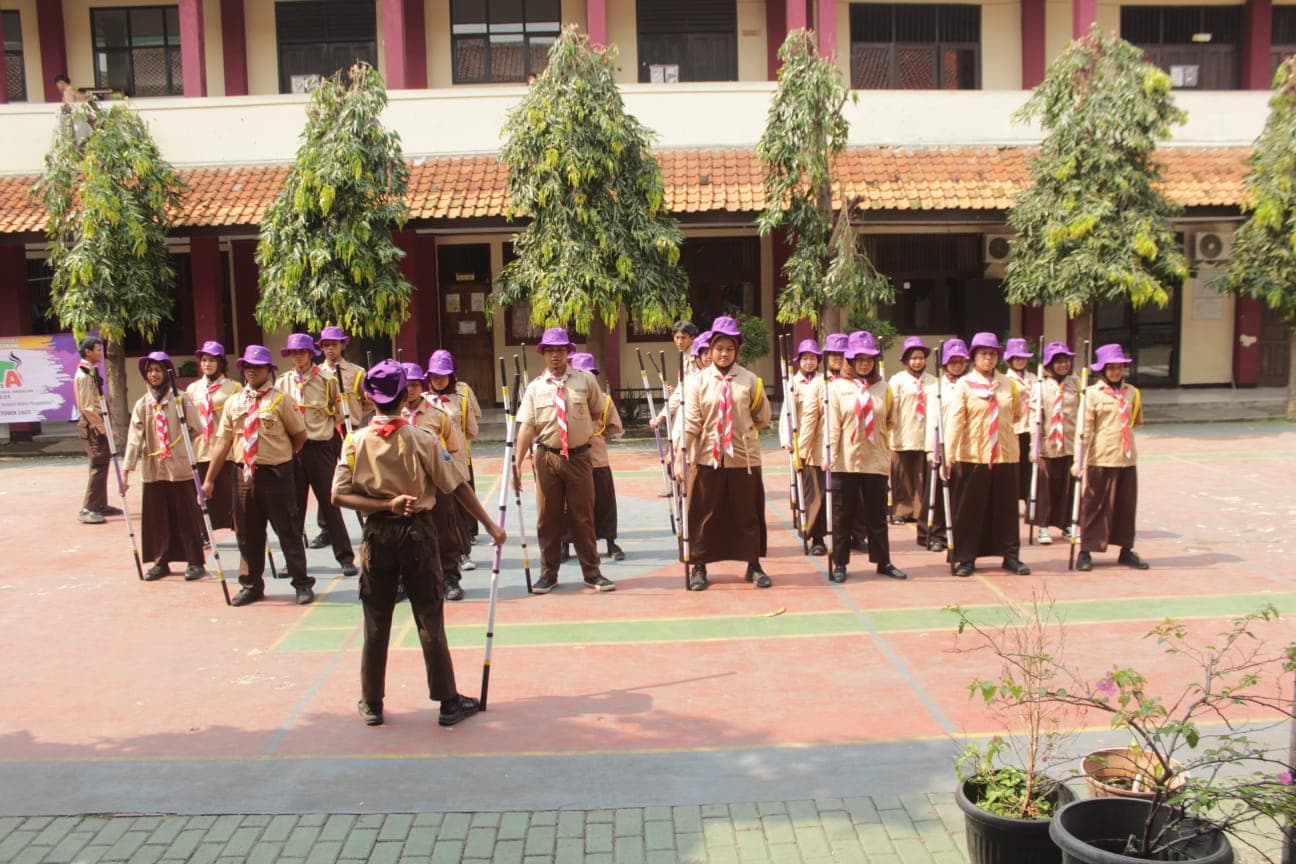 Anggota pramuka bertopi ungu berbaris rapi memegang tongkat di lapangan sekolah saat kegiatan latihan.