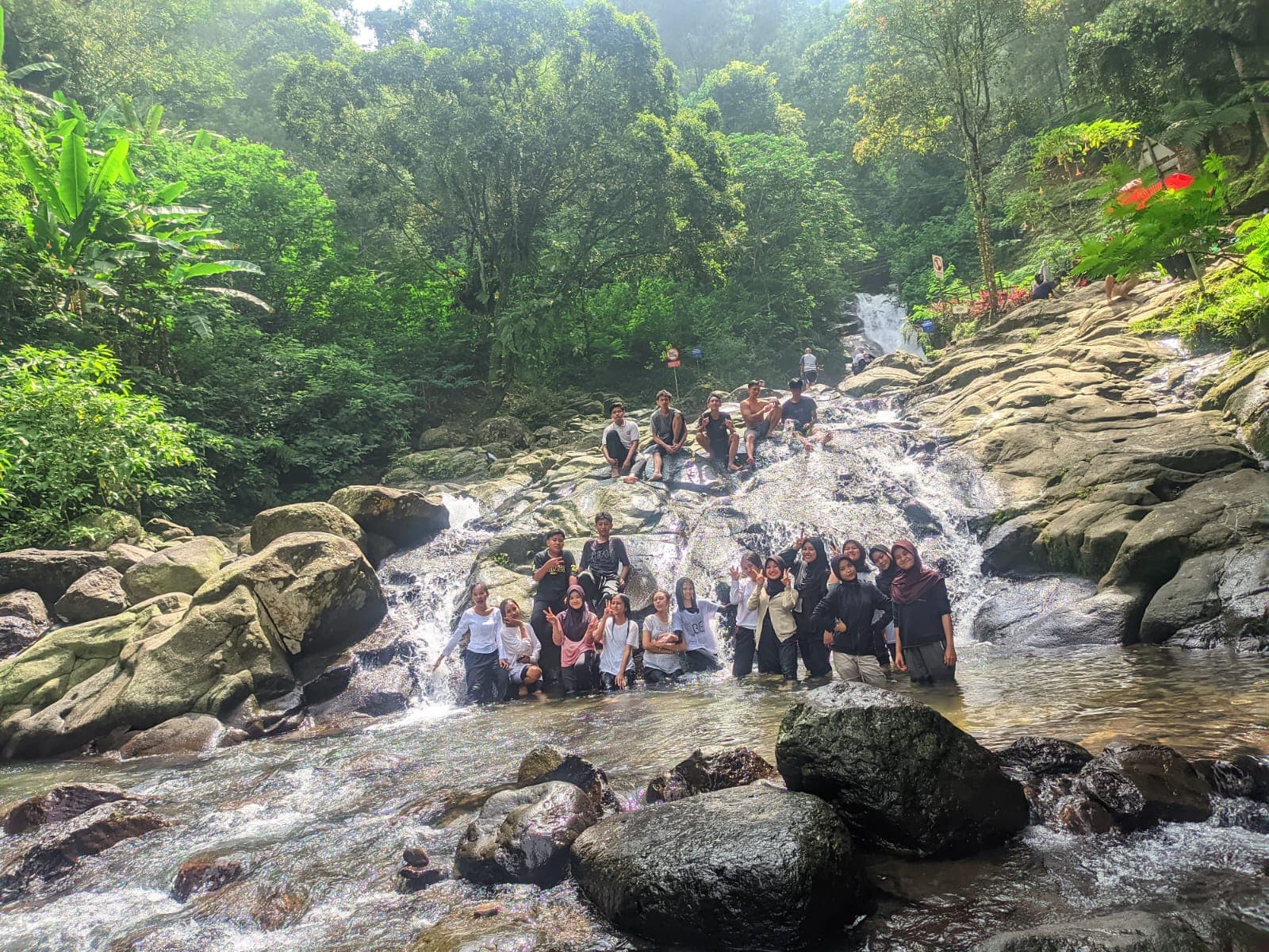 Sekelompok orang berpose di air terjun berbatu yang dikelilingi oleh pepohonan hijau yang rimbun.