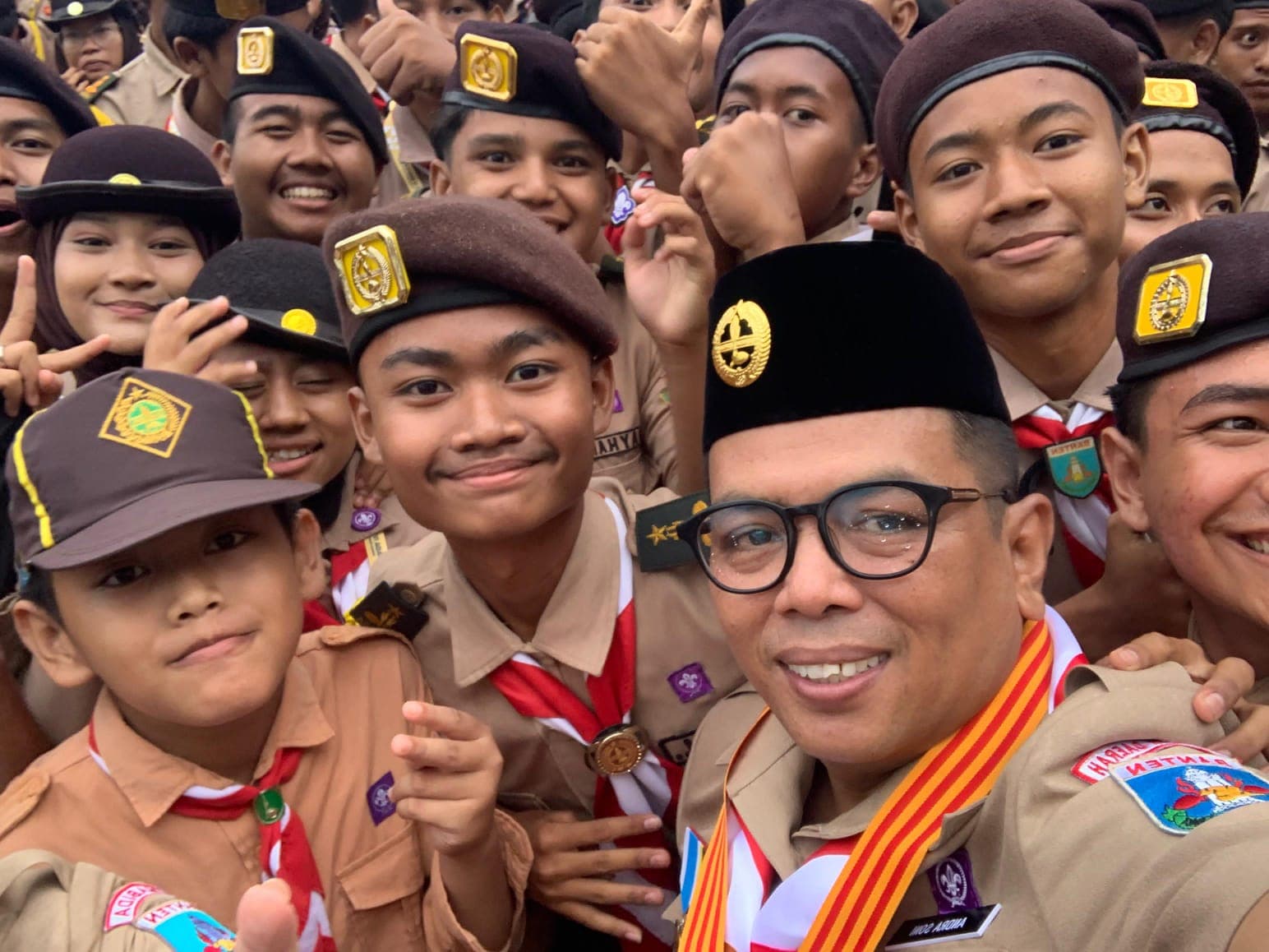 Sekelompok anggota pramuka Indonesia tersenyum ceria bersama seorang pria berkacamata dan mengenakan peci hitam.