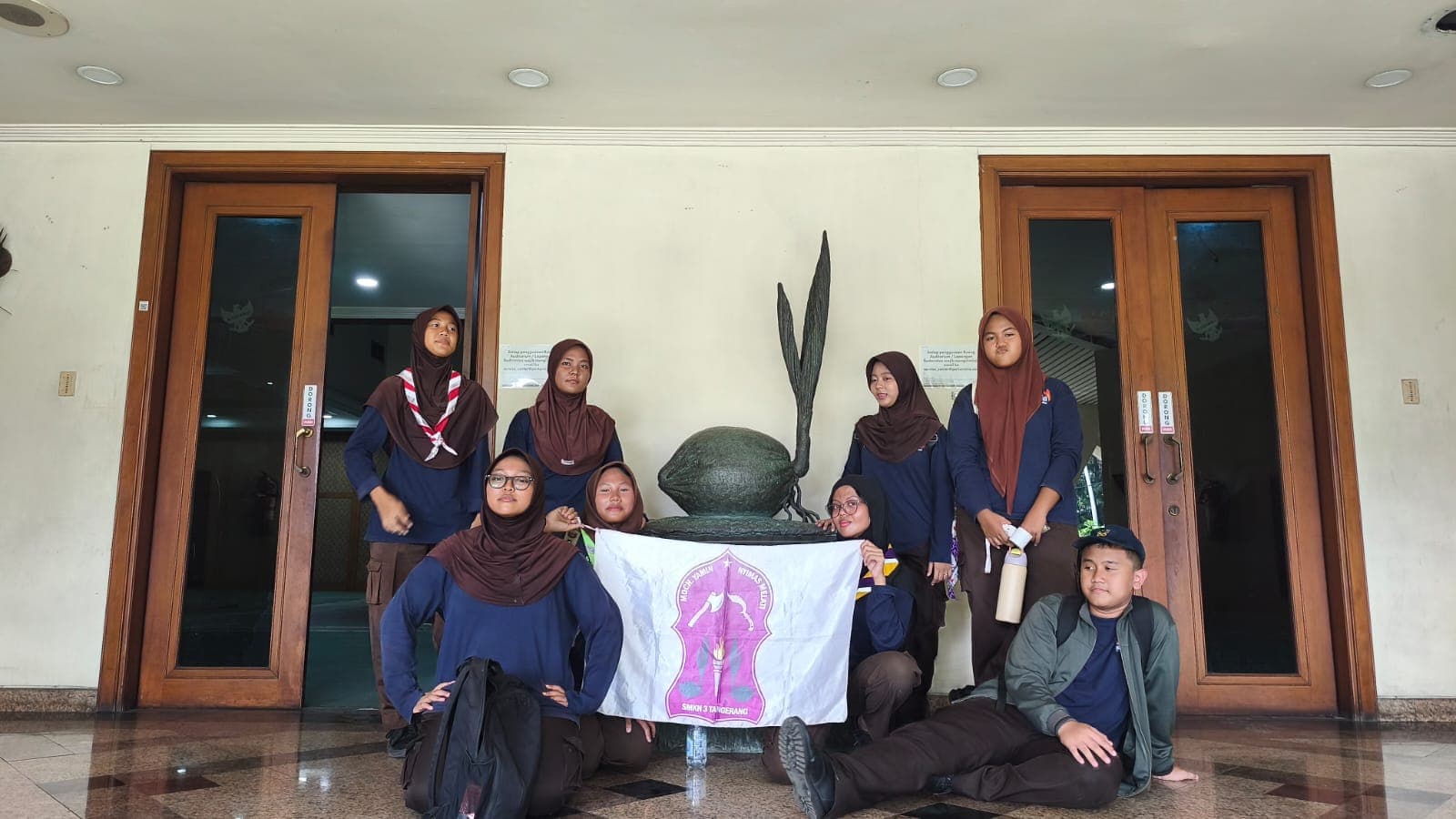 Sekelompok siswa pramuka berpose memegang bendera di depan patung tunas kelapa sebuah gedung.
