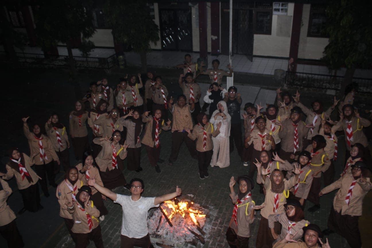 Sekelompok anggota pramuka berpose ceria di sekitar api unggun yang menyala terang pada malam hari.