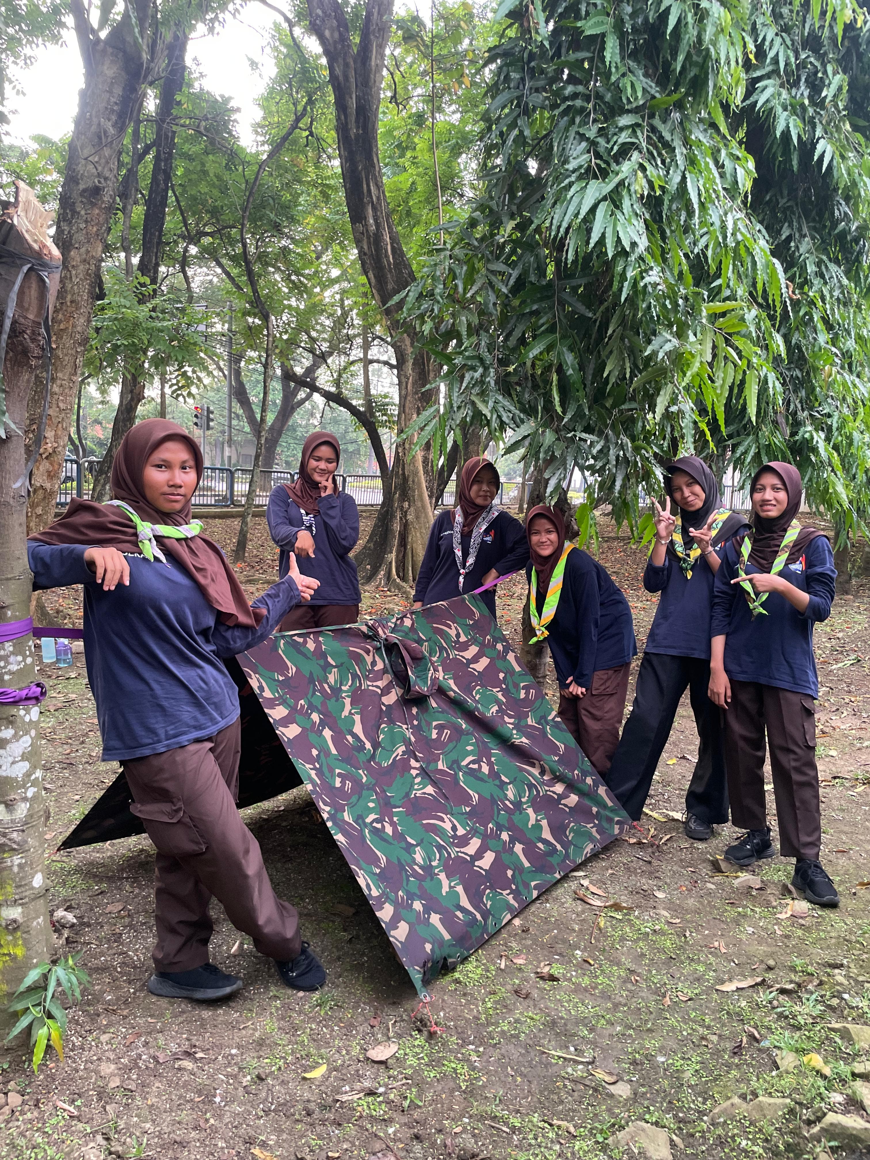 Sekelompok pramuka perempuan berhijab berpose di sekitar tenda bivak bermotif loreng di bawah pepohonan.
