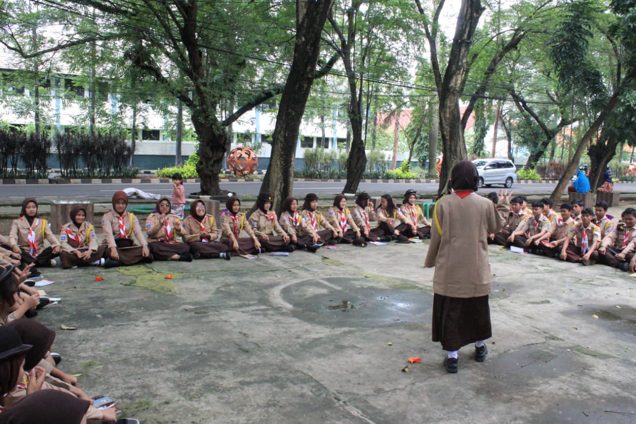 Sekelompok siswa berseragam pramuka duduk melingkar di luar ruangan mendengarkan instruksi dari seorang pemimpin.