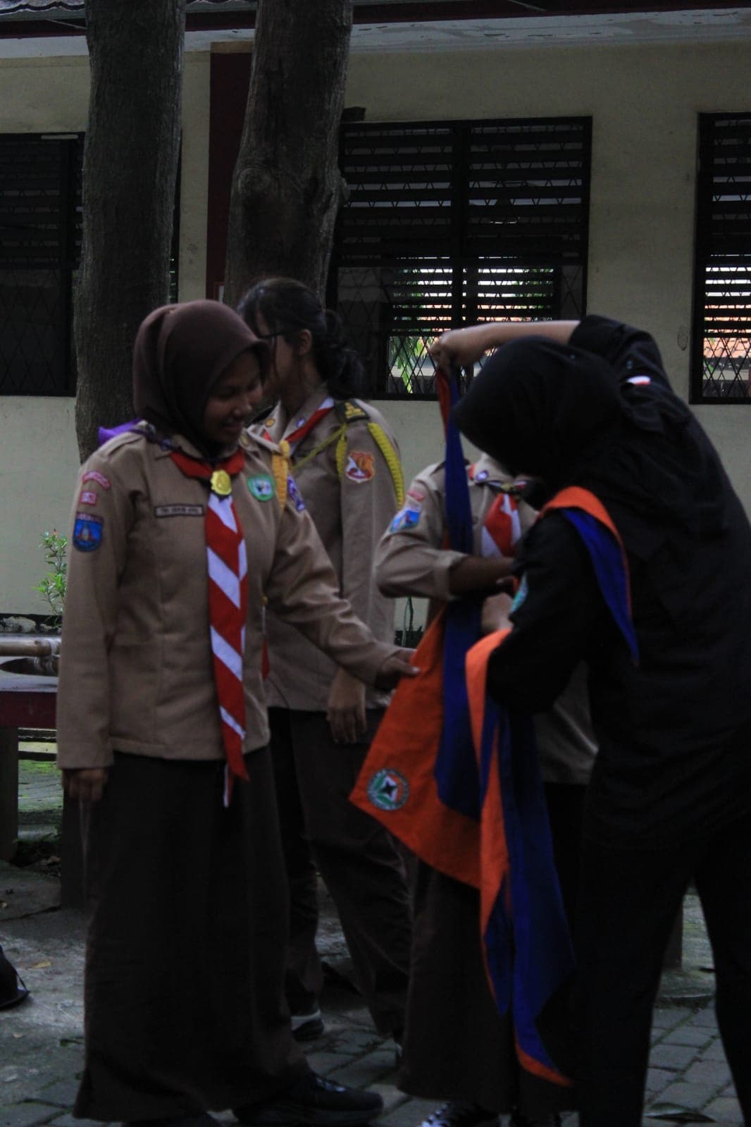 Siswa pramuka berhijab tersenyum saat rekannya menyiapkan kain bendera berwarna oranye dan biru.