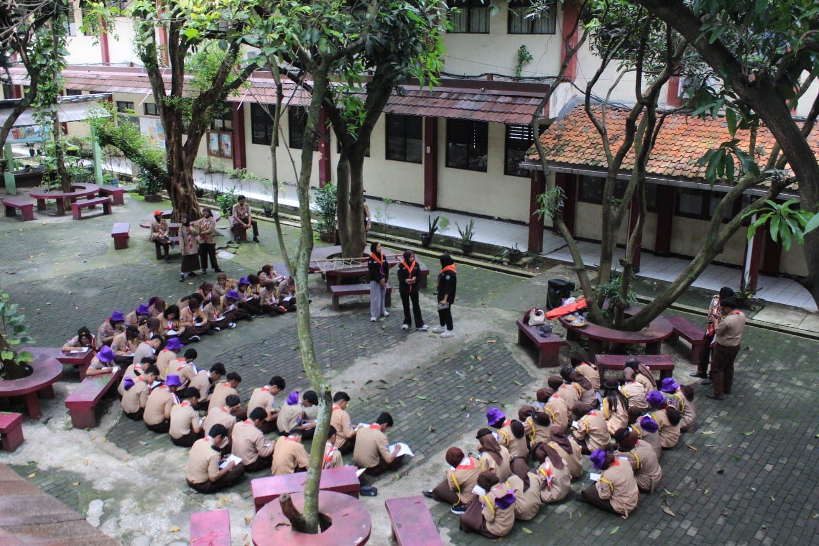 Sekelompok siswa pramuka duduk melingkar di halaman sekolah yang rindang mendengarkan instruksi pembina.