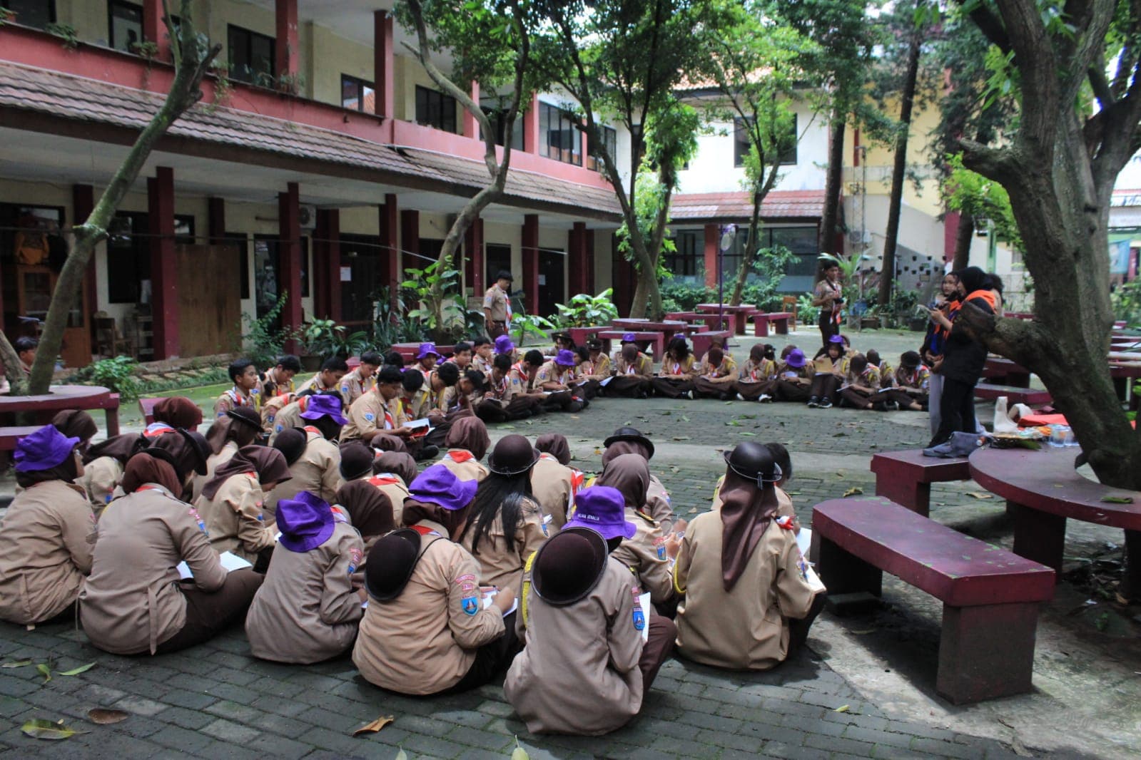 Sekelompok siswa berseragam pramuka duduk melingkar di halaman sekolah yang rindang untuk kegiatan luar kelas.