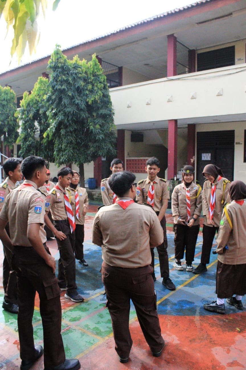 Sekelompok pramuka berdiri melingkar di lapangan sekolah yang berwarna-warni di depan gedung sekolah.