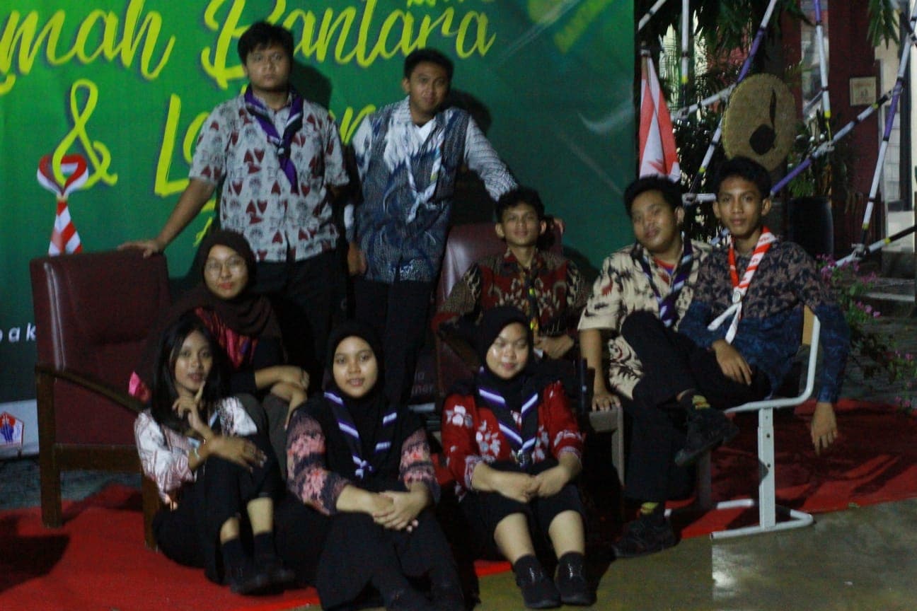 Sekelompok siswa mengenakan batik dan hasduk pramuka berpose bersama di depan spanduk acara hijau.
