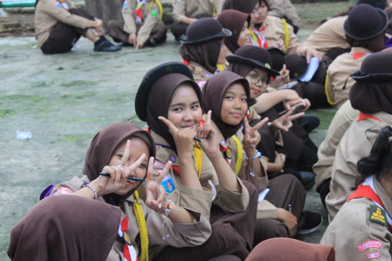 Siswi Pramuka berhijab cokelat tersenyum dan berpose dua jari saat berkumpul di luar ruangan.