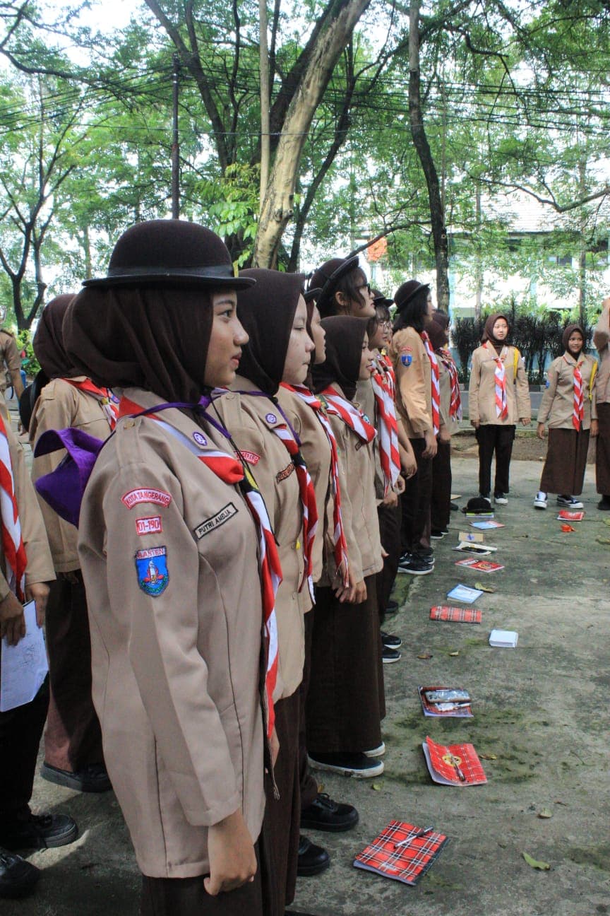 Sekelompok siswi pramuka berhijab berdiri berbaris rapi mengenakan seragam lengkap di bawah pepohonan rindang.