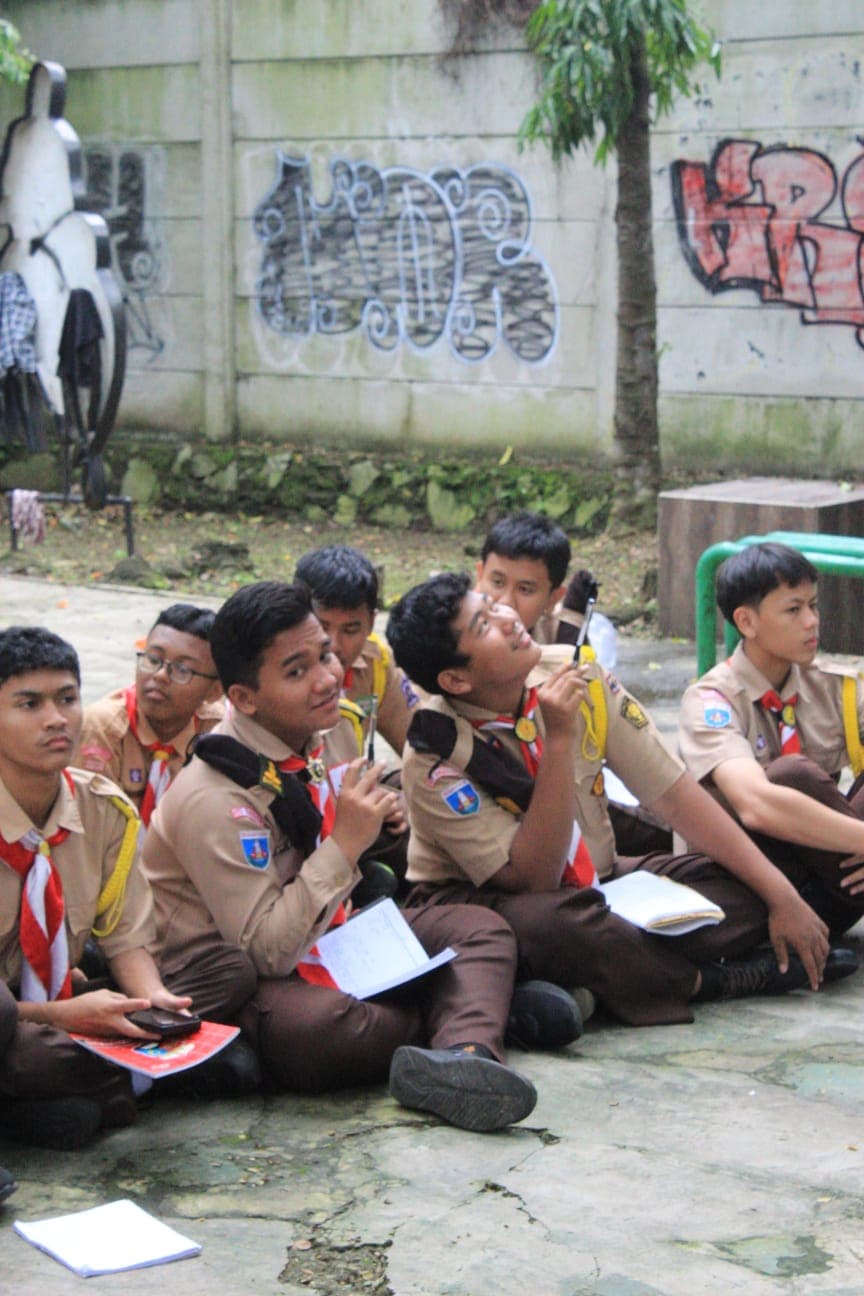 Sekelompok anggota pramuka laki-laki duduk di tanah sambil memegang buku catatan dan pulpen.